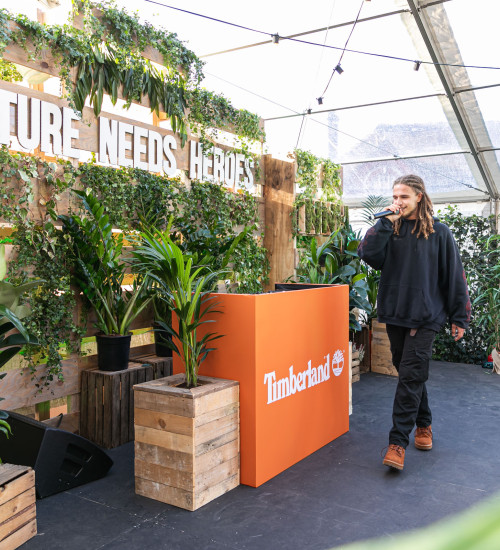 Young man with microphone surrounded by planted and walking past a DJ booth with the Timberland logo on it.